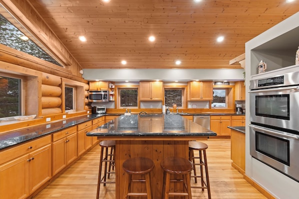 Una cocina moderna con armarios de madera, una isla central con taburetes, electrodomésticos de acero inoxidable y ventanas amplias.