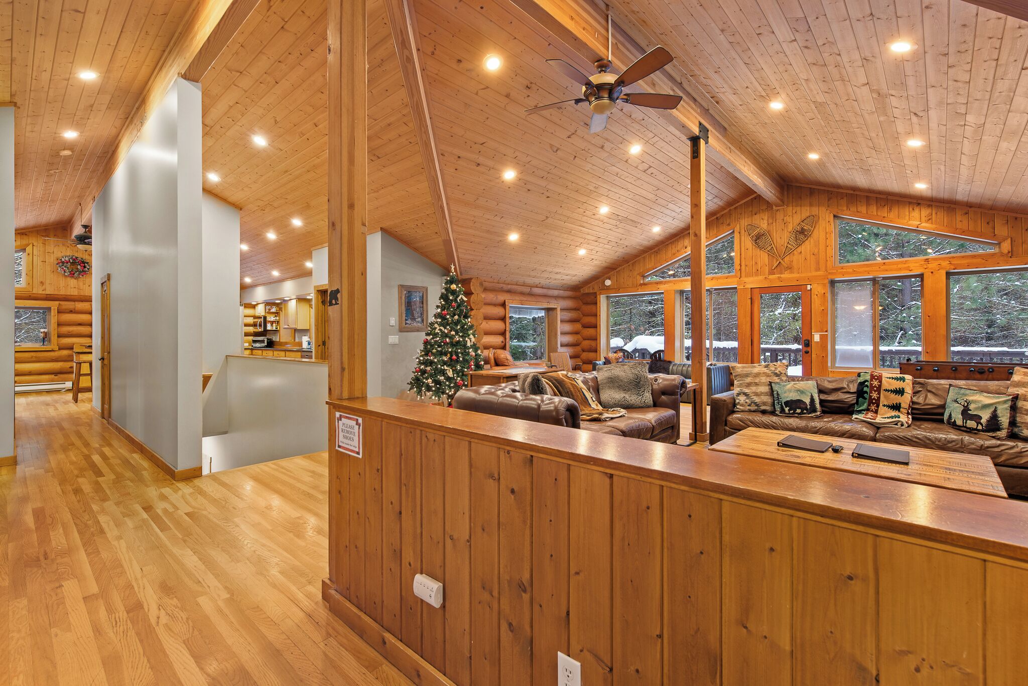 Interior de uma cabana de madeira com teto rústico, área de estar central decorada com uma árvore de Natal e grandes janelas.