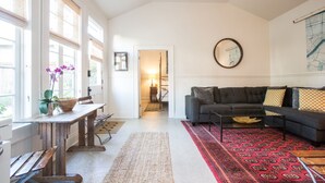 A spacious living room with a dark grey sofa, a wooden coffee table, and a red patterned rug. There is a large window with blinds, a ceiling fan, and a framed map on the wall. - Historic Uptown Cottage - 1 mile to Tulane & Loyola (New Orleans)