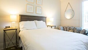 A neatly arranged bedroom with a large bed, two bedside tables with lamps, a round mirror on the wall, and a patterned sofa. - Historic Uptown Cottage - 1 mile to Tulane & Loyola (New Orleans)