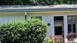 A white building with a covered patio, two green chairs, a table, and a garden with various plants. - Historic Uptown Cottage - 1 mile to Tulane & Loyola (New Orleans)