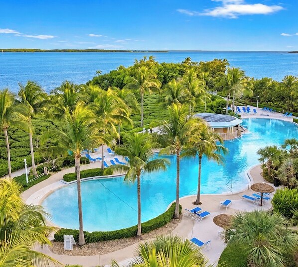 Piscina de resort con palmeras y sillas de descanso, rodeada de exuberante vegetación y vista al mar.