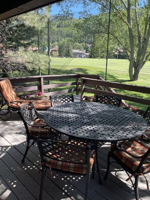 Un patio techado al aire libre con una mesa metálica redonda y sillas, un banco de madera y vistas a un césped cuidado y árboles.