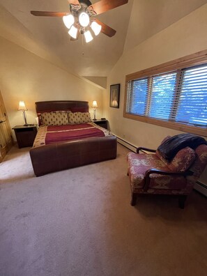 Un dormitorio con cama, dos mesitas de noche con lámparas, ventilador de techo, ventana con persianas y un sillón estampado.