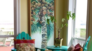 Une pièce avec des fauteuils de couleur turquoise, un coussin à motifs floraux, un tableau représentant un palmier et une vue par la fenêtre.