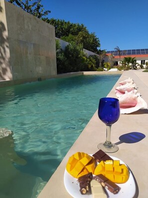 A poolside setting with a clear pool, a glass of blue beverage, and a plate of fruit and chocolate.