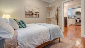 Een slaapkamer met een bed, nachtkastje, lamp, kapstok en een schilderij van een stadsgezicht.