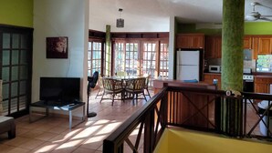 Een ruime woonkamer met een televisie, eettafel en stoelen, een keuken met kastjes en apparatuur, en grote ramen die voor veel daglicht zorgen.