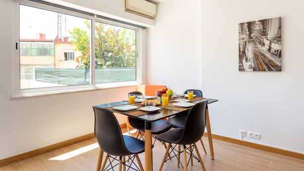A dining area with a table set for four, a window with a view of buildings and trees, and a wall-mounted air conditioner. - Graça House - Apartment T1 (Lisboa)