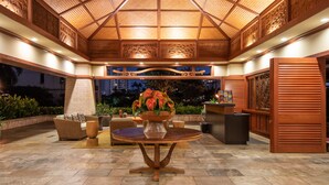 An outdoor patio area with a wooden ceiling, a round table with a vase of flowers, wicker furniture, and a brick structure in the background.