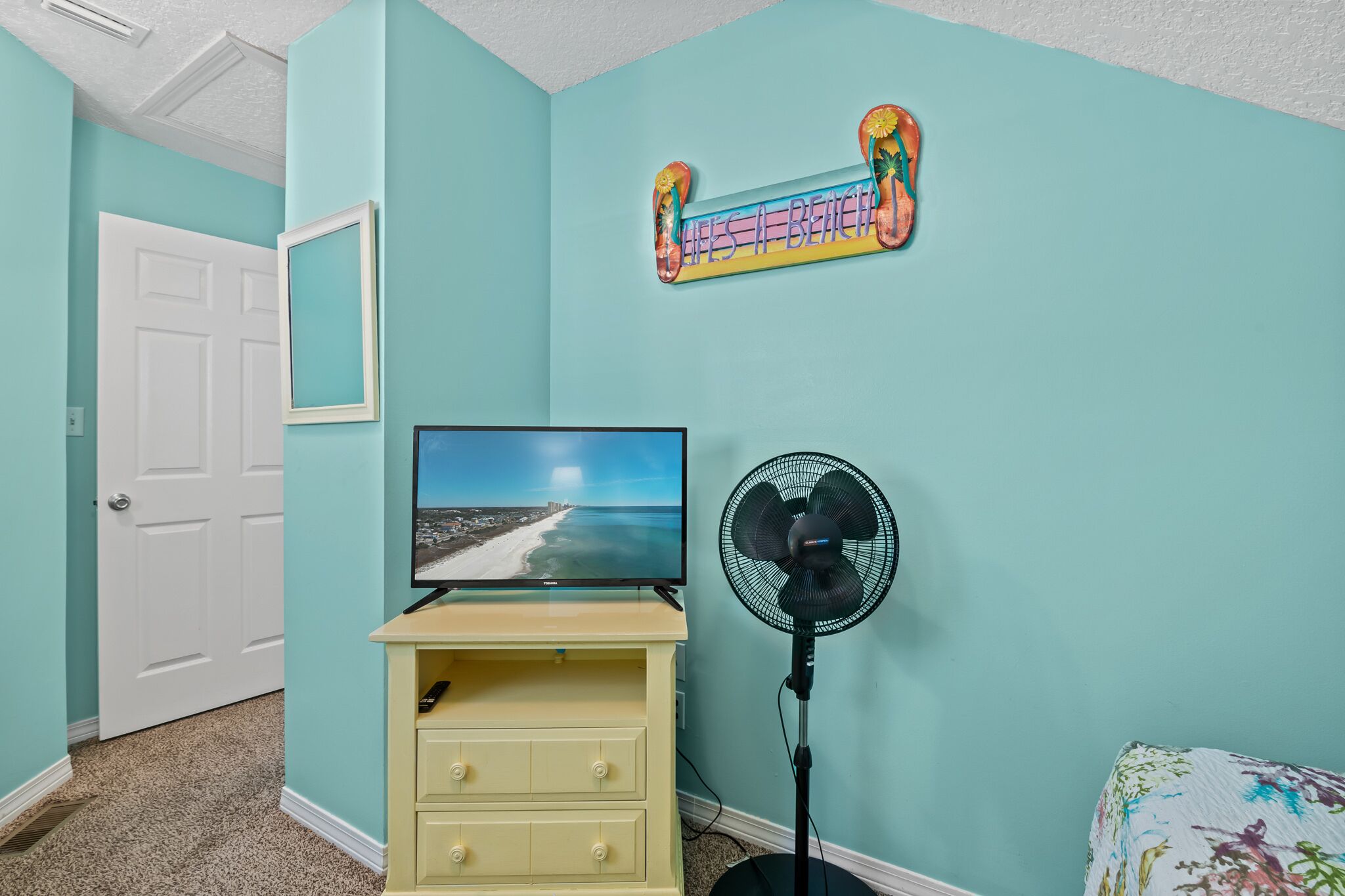 Quarto com paredes verde-água, porta branca, televisão sobre uma cômoda amarela, ventilador e placa com tema praiano.