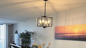 A dining area with a glass table surrounded by wicker chairs, a hanging light fixture, and a painting on the wall. - 5 Star Marco Island Beachfront Condo – Limited-Time Deals Available! (Marco Island)