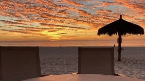 A table with a mug, overlooking a beach at sunset with a thatched umbrella. - Ocean view, one bedroom, steps to the pool and beach.  Pride in ownership. (San Jose del Cabo)
