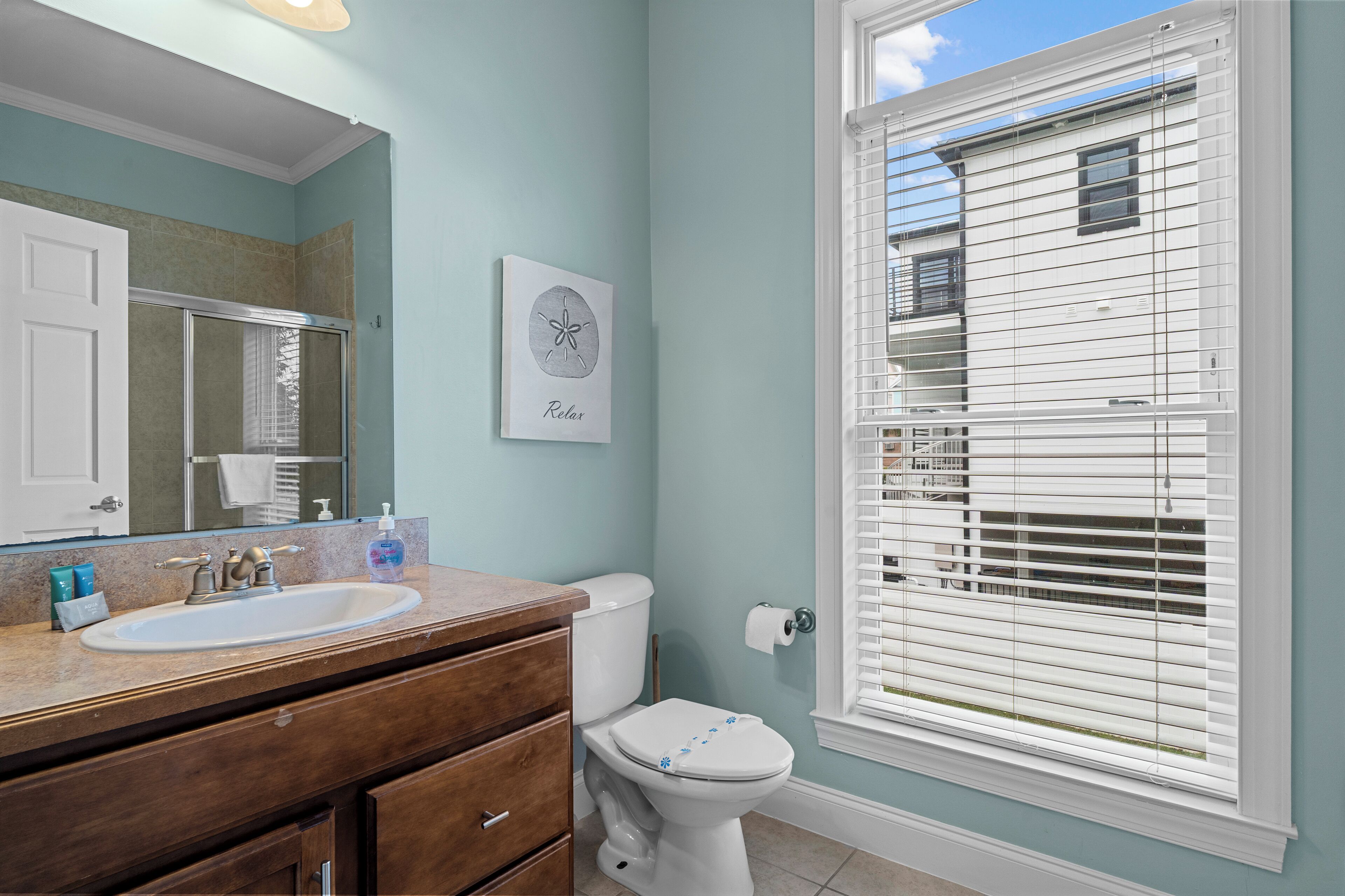 A bathroom with a white sink, wooden vanity, and a toilet. There is a window with blinds and a framed picture on the wall.