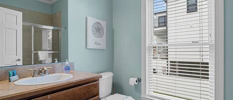 A bathroom with a white sink, wooden vanity, and a toilet. There is a window with blinds and a framed picture on the wall.