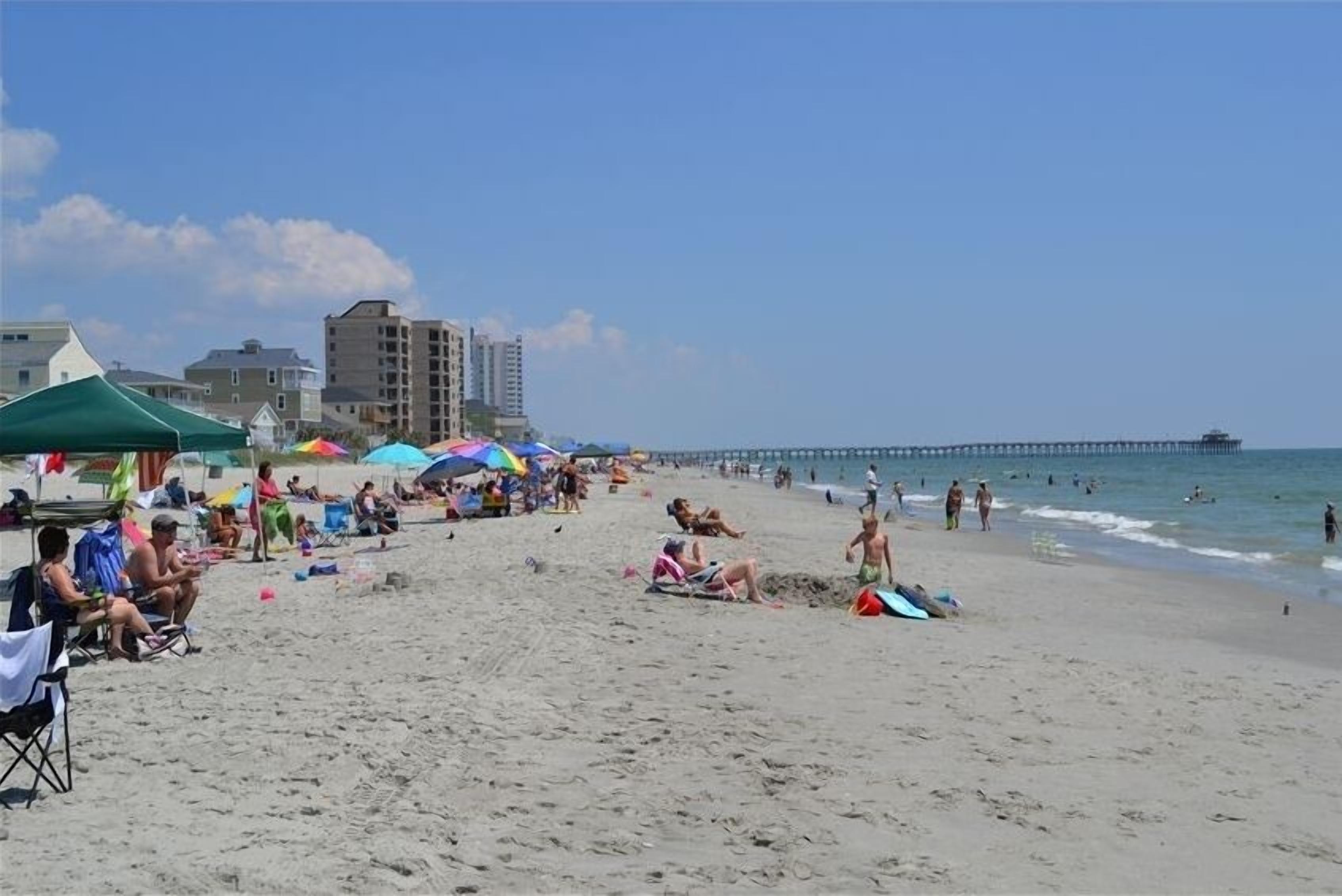 Une plage où des gens se prélassent au soleil et se baignent, une jetée en arrière-plan et des bâtiments à proximité.