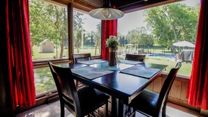 Un coin repas avec une table dressée pour quatre personnes, de grandes fenêtres ornées de rideaux rouges et une vue sur un jardin avec une piscine et des arbres.