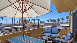 A balcony with wicker furniture, a large umbrella, and a view of palm trees and buildings. - OCEANSIDE BEACH located by pier and marina! Ocean Views! Walk to Village! (Oceanside)