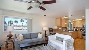 A modern living room with a grey sofa, a white sofa, and a wooden coffee table. There is a ceiling fan, a window with a view of palm trees, and a kitchen area with wooden cabinets and a stainless steel refrigerator. - OCEANSIDE BEACH located by pier and marina! Ocean Views! Walk to Village! (Oceanside)