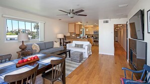 A modern living room with a dining area, kitchen, and a large TV mounted on the wall. - OCEANSIDE BEACH located by pier and marina! Ocean Views! Walk to Village! (Oceanside)