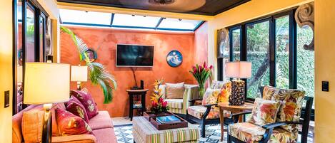 Un salon confortable avec un canapé, des fauteuils et une table basse. Un téléviseur est fixé au mur et un ventilateur de plafond est installé.