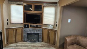 A living room with a stone fireplace, wooden cabinets, and a flat-screen TV. - Catalina Park Model, in RV Ballard RV Park  (Thompson)