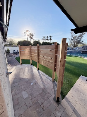 A patio area with a wooden fence, a brick pathway, and a view of a swimming pool and palm trees. - Huge Heated Pool, Rope Swing, Slide, Spa, pickle ball on 1 acre!!!! (Mesa)
