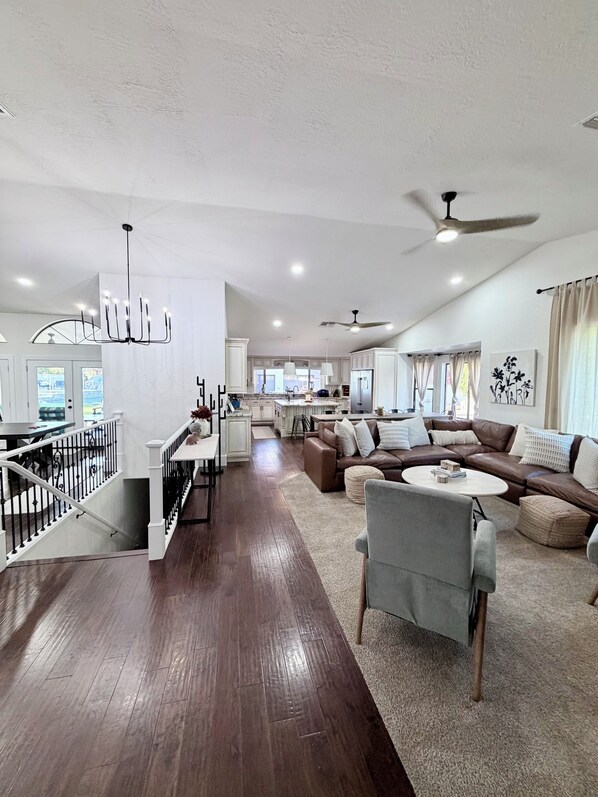 A spacious living area with a dark wooden floor, a brown leather sofa, a grey armchair, and a dining area with a chandelier.