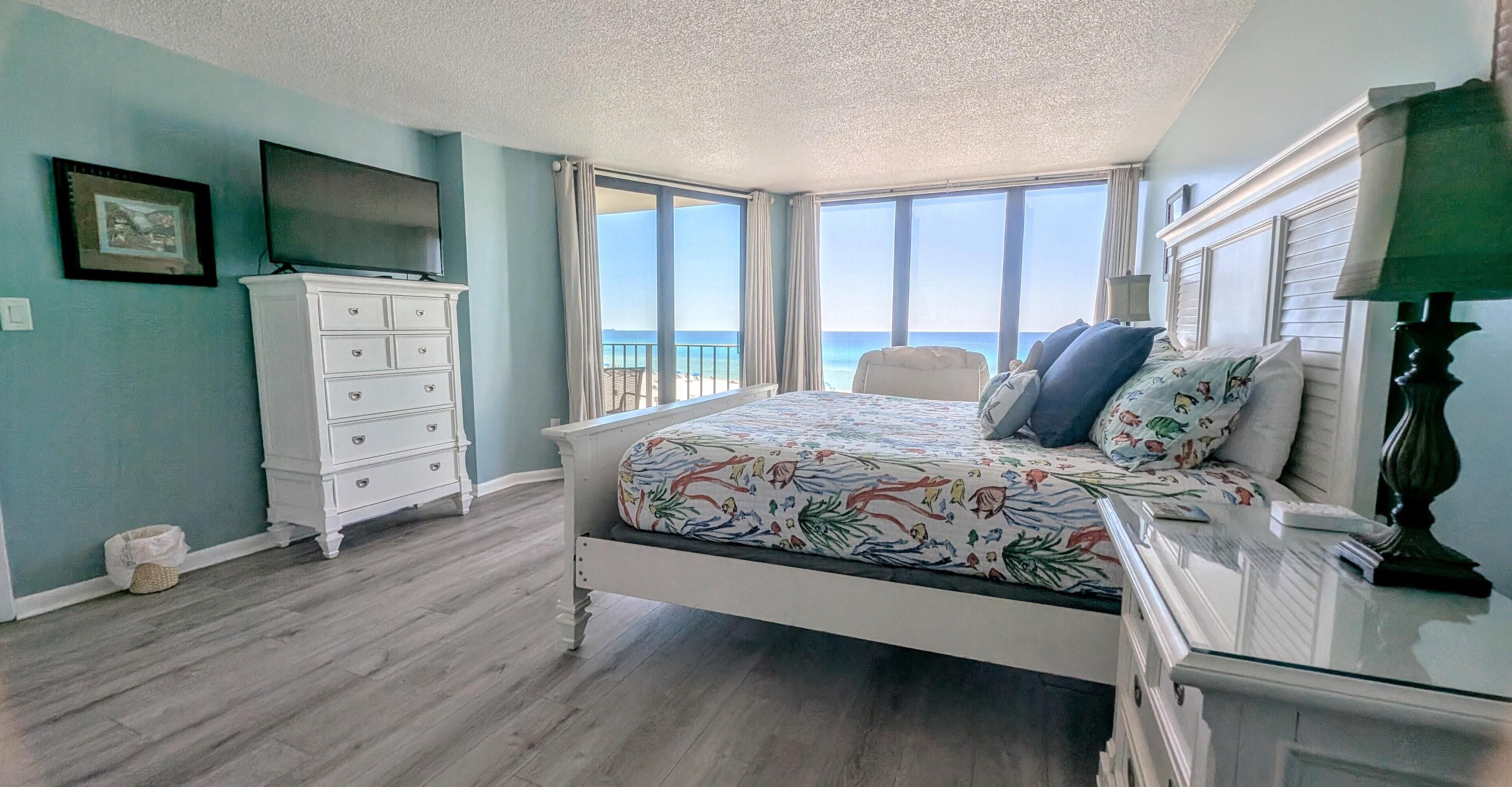 A bedroom with a bed, dresser, TV, and a view of the ocean.
