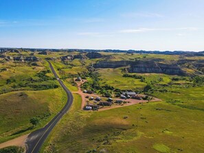 Paisaje rural con un camino serpenteante, un pequeño asentamiento con casas y vehículos, y colinas onduladas al fondo.