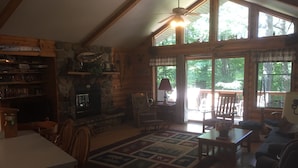 L’intérieur chaleureux d’un chalet avec une cheminée en pierre, des meubles en bois et de grandes fenêtres offrant une vue sur le paysage extérieur.
