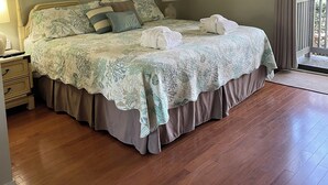 Ein Schlafzimmer mit einem großen Bett, zwei gerahmten Pflanzenbildern an der Wand, einem Nachttisch mit Lampe und Blick ins Freie durch ein Fenster.