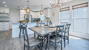 A modern kitchen with white cabinetry, a central island, and a dining area with metal chairs and a wooden table. - Beautifully Remodeled - Short Walk to Beach! (Gulf Shores)