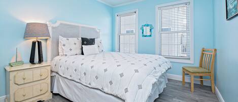Quarto com cama, mesa de cabeceira de madeira, cadeira, ventilador de teto e duas janelas com persianas.