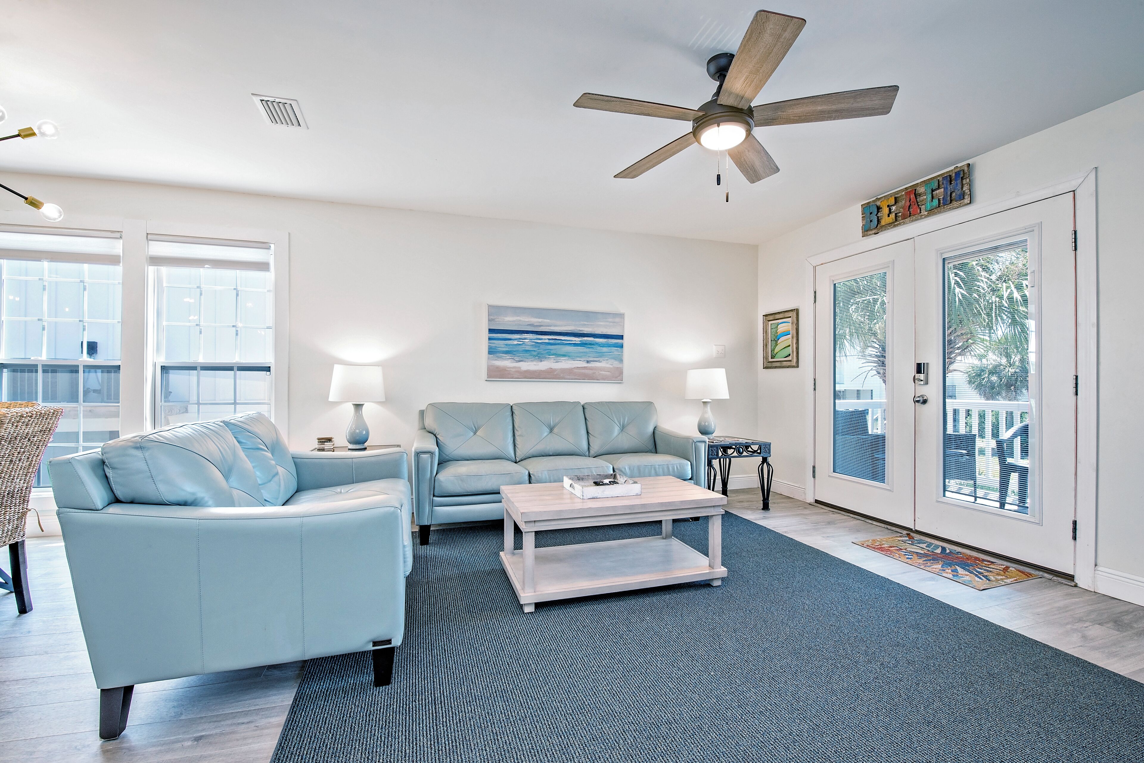 A living room with a light blue sofa, a coffee table, and a ceiling fan. There is a painting on the wall and a door with glass panels.