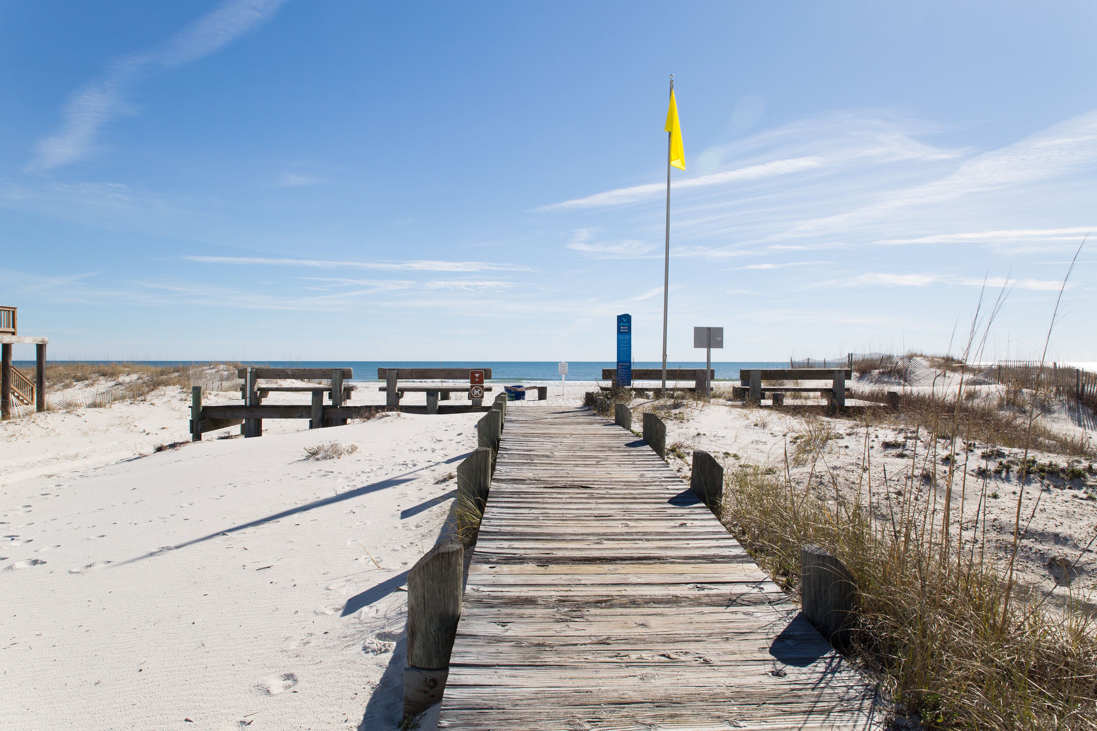 A wooden boardwalk leading to a sandy beach with benches and a flag.