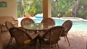 A glass-top table surrounded by wicker chairs in a patio area with a view of a pool and tropical plants. - Villa Salva, Your Home Away From Home! (Cape Coral)