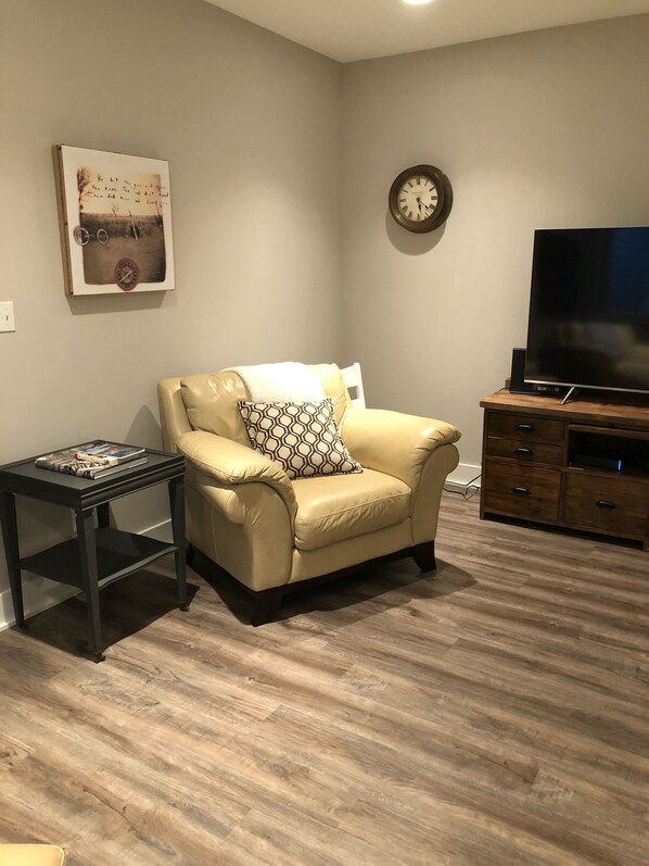 A living room with a beige leather armchair, a small side table with a book, a clock on the wall, and a TV on a wooden cabinet.