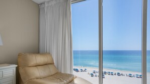 A room with a large window overlooking a beach, a beige leather chair, and a white bedside table.