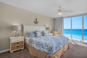 A bedroom with a large bed, two bedside tables, a ceiling fan, and a view of the ocean.