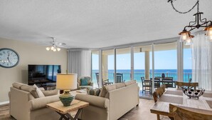 A living room with a sofa, TV, and a table with a lamp. There is a balcony with a view of the ocean.