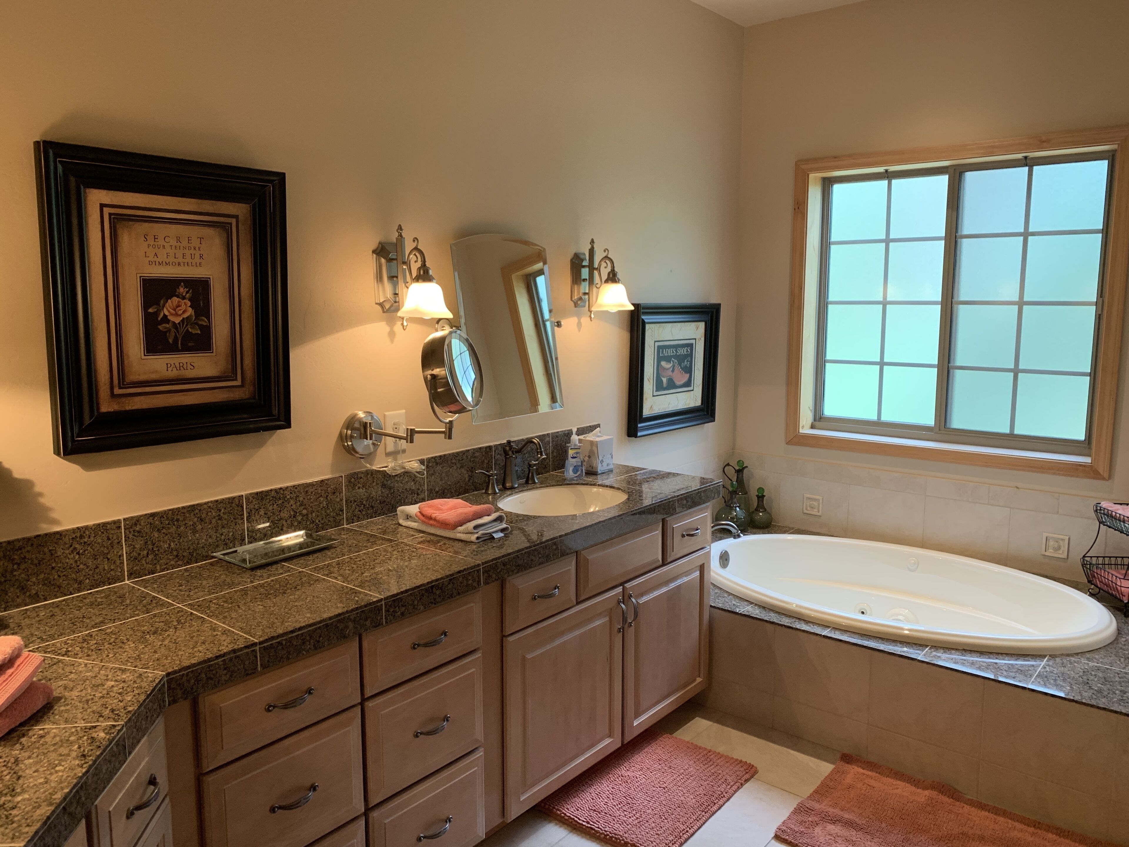 A bathroom with a large bathtub, a sink, and a vanity with drawers. There are framed pictures on the wall and a window providing natural light.