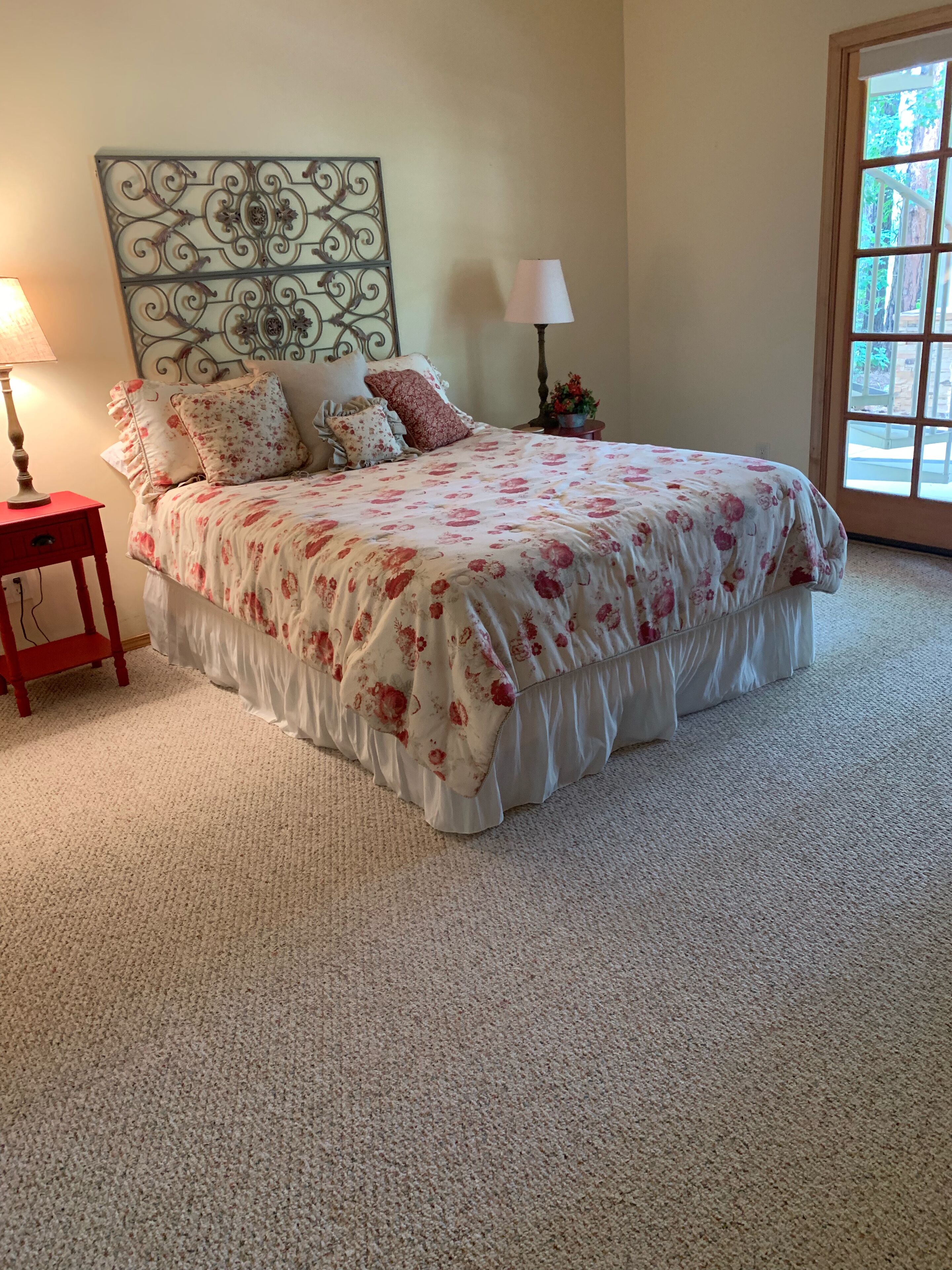 A bedroom with a large bed, a headboard with a decorative metal design, two bedside tables with lamps, and a window with a view of greenery.