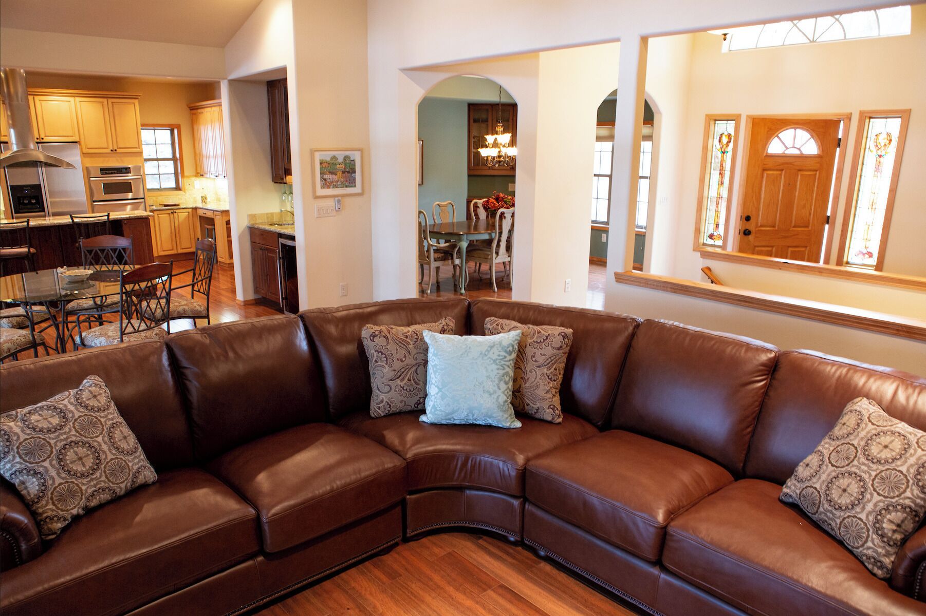 A spacious living room with a brown leather sectional sofa, wooden flooring, and a view into the kitchen area.