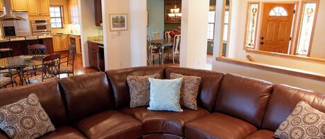 A spacious living room with a brown leather sectional sofa, wooden flooring, and a view into the kitchen area.