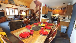 A dining area with a wooden table set for six, red plates, and matching cups. A chandelier hangs from the ceiling, and a staircase leads to an upper level.