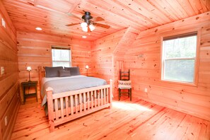 A wooden cabin bedroom with a bed, two windows, a ceiling fan, and a chair.