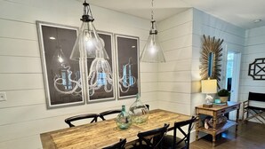 Un coin repas avec une table en bois, des chaises noires et des luminaires suspendus. Des œuvres d’art encadrées ornent le mur, et une petite table d’appoint, sur laquelle se trouvent une lampe et des objets décoratifs, complète l’ensemble.