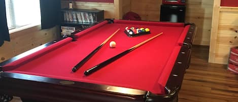 Una mesa de billar con bolas y tacos, un microondas y un cartel que dice 'Juegos de Mesa' en una habitación con piso de madera y una ventana.