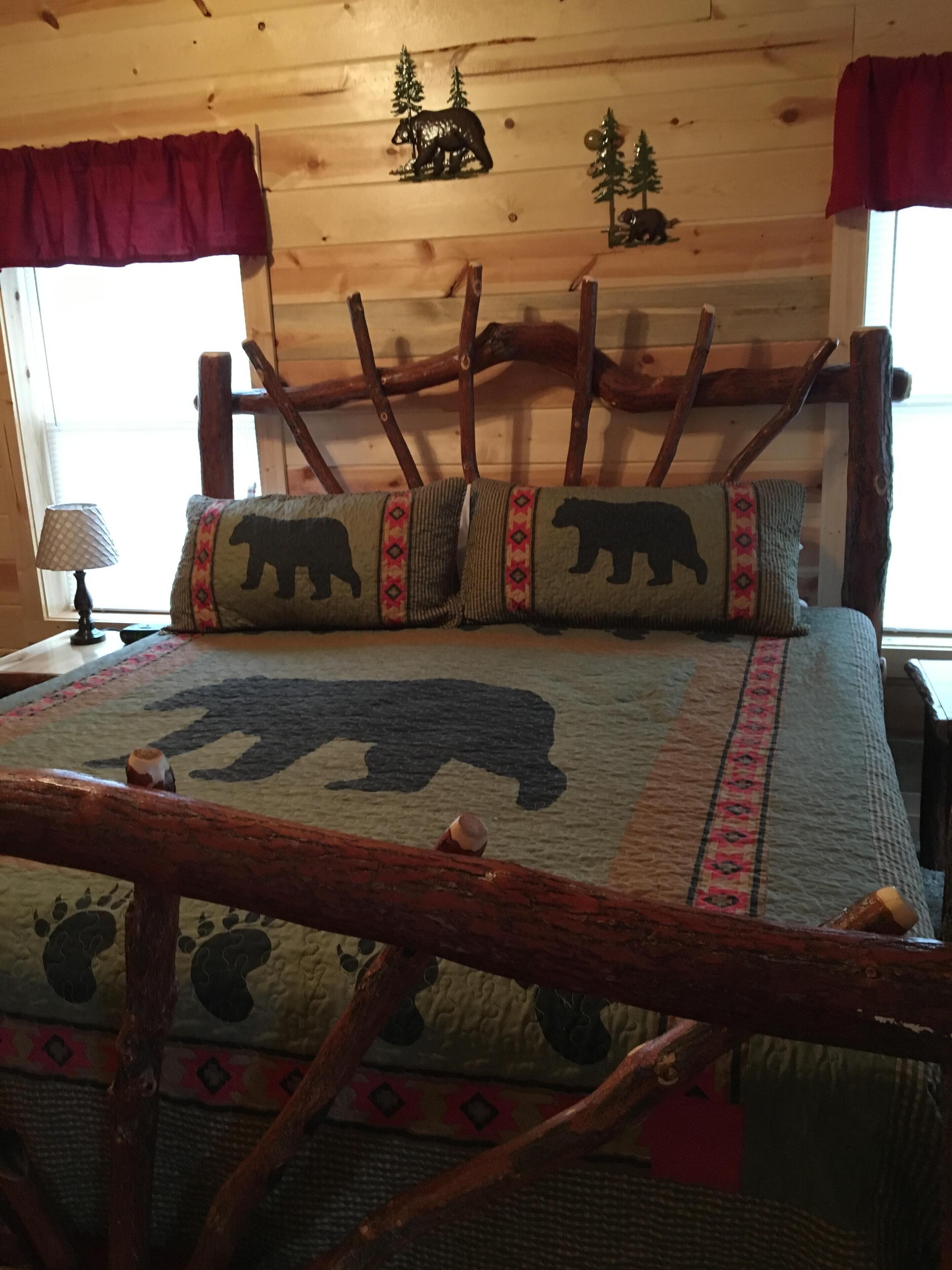 Interior de una cabaña de madera con una cama que incluye colcha con motivo de oso, cabecera de madera y ventana con vistas a árboles.